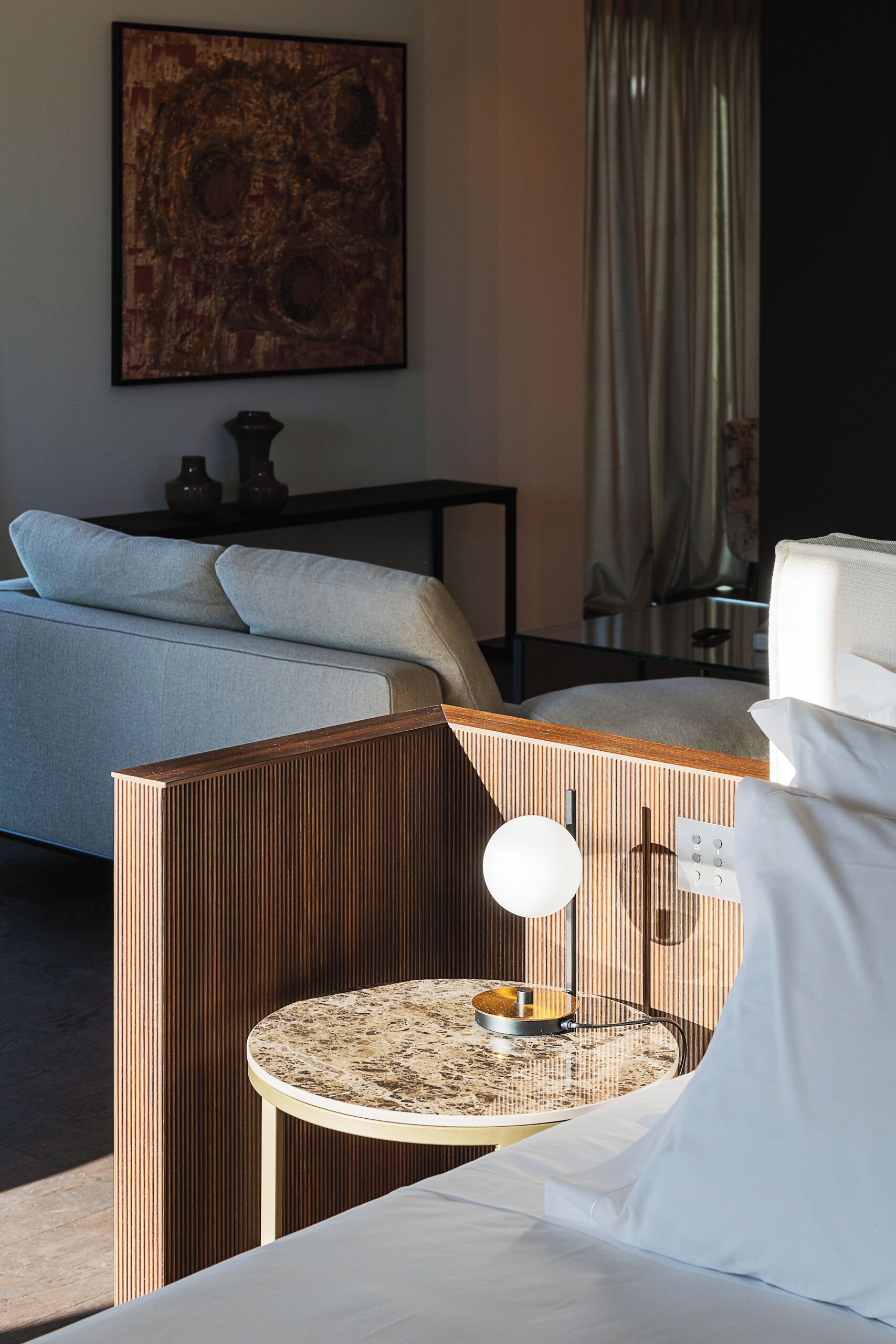 A hotel room with a bed, a bedside table with a lamp, a small round table with a bowl, a painting on the wall, and a curtain.