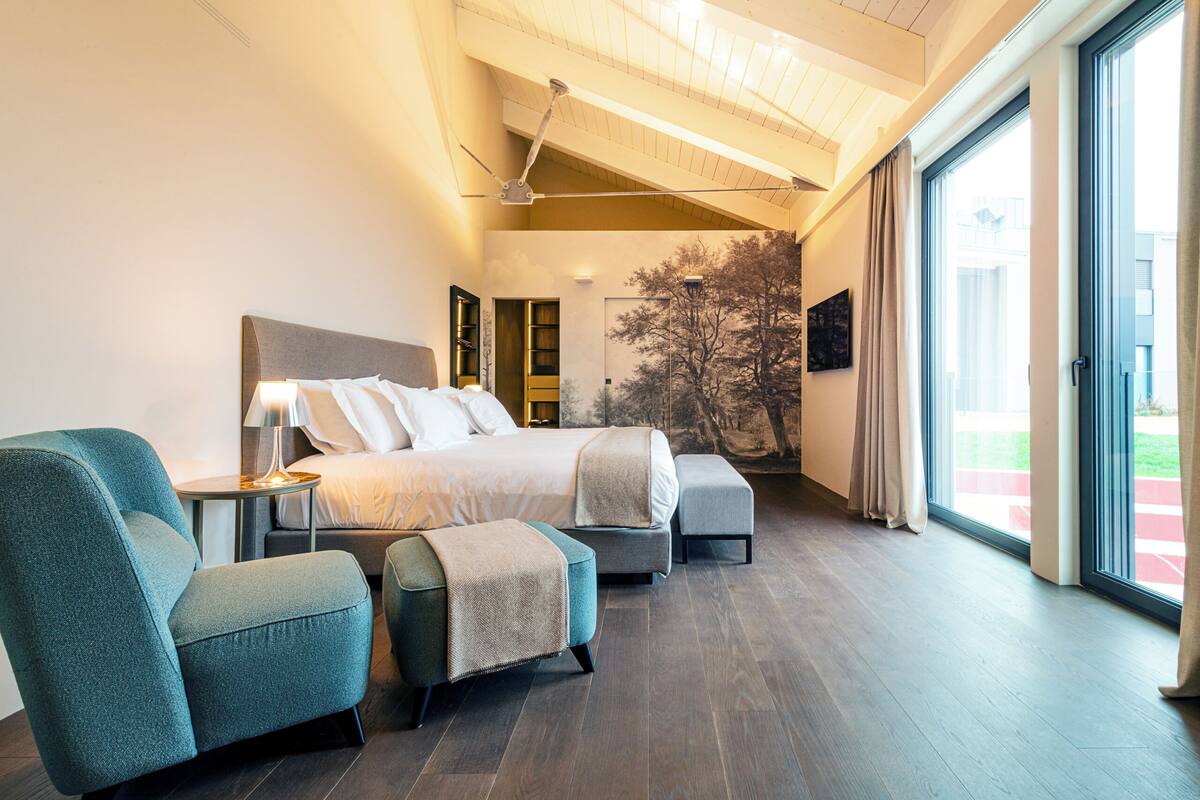 A modern bedroom with a large bed, two armchairs, a small table, and a large window with a view of the outdoors.