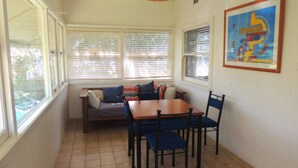 A room with a wooden table and chairs, a sofa with patterned cushions, and a window with blinds.