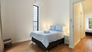 A bedroom with a bed, bedside tables, a window with blinds, and a door leading to another room.