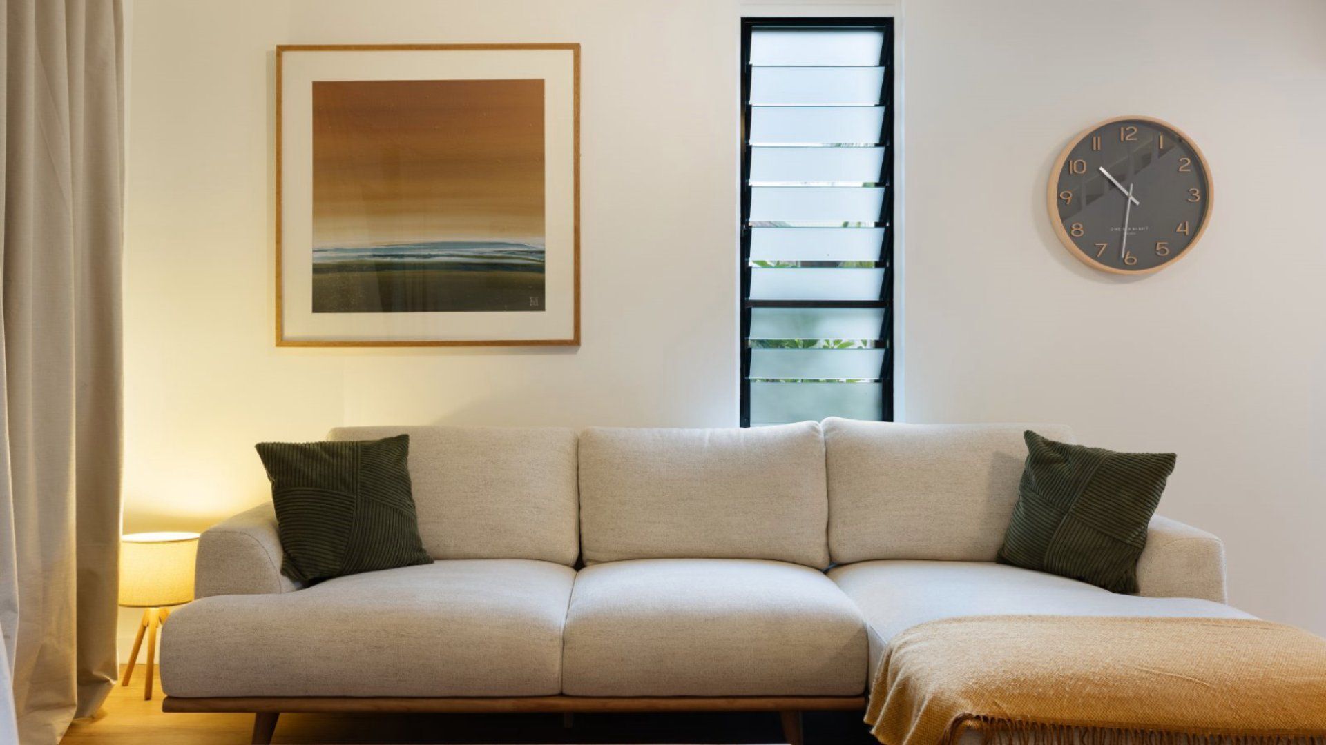 A living room with a sofa, a framed picture, a window with blinds, and a wall clock.