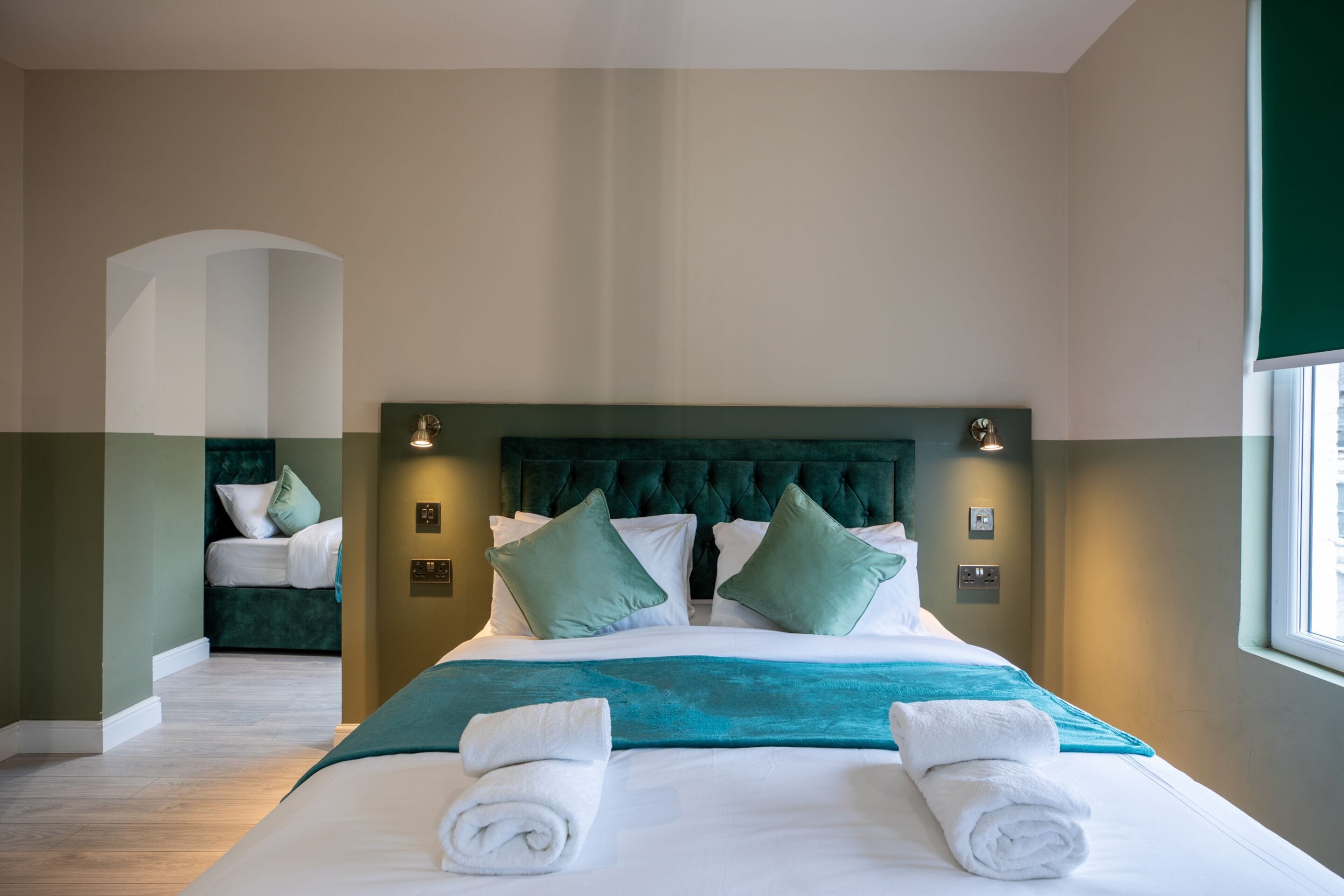 A hotel room with a large bed, green and white bedding, two pillows, and a teal throw blanket.