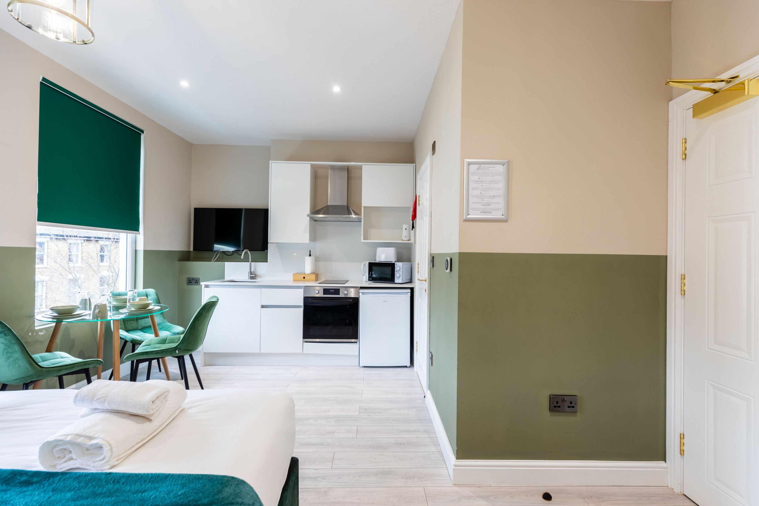 A compact hotel room with a kitchenette, a dining area with a table and chairs, and a bed with a teal blanket.