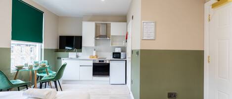A compact hotel room with a kitchenette, a dining area with a table and chairs, and a bed with a teal blanket.