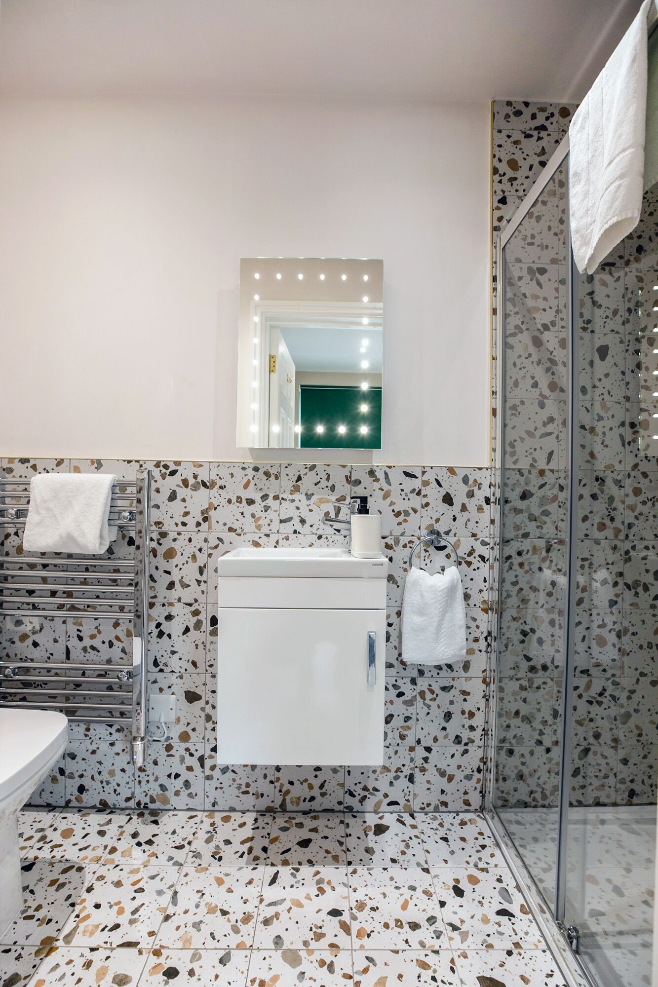 Un baño con un lavamanos blanco, un espejo con iluminación y una ducha con puerta de vidrio, además de suelo de terrazo.