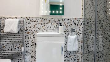 Un baño con un lavamanos blanco, un espejo con iluminación y una ducha con puerta de vidrio, además de suelo de terrazo.