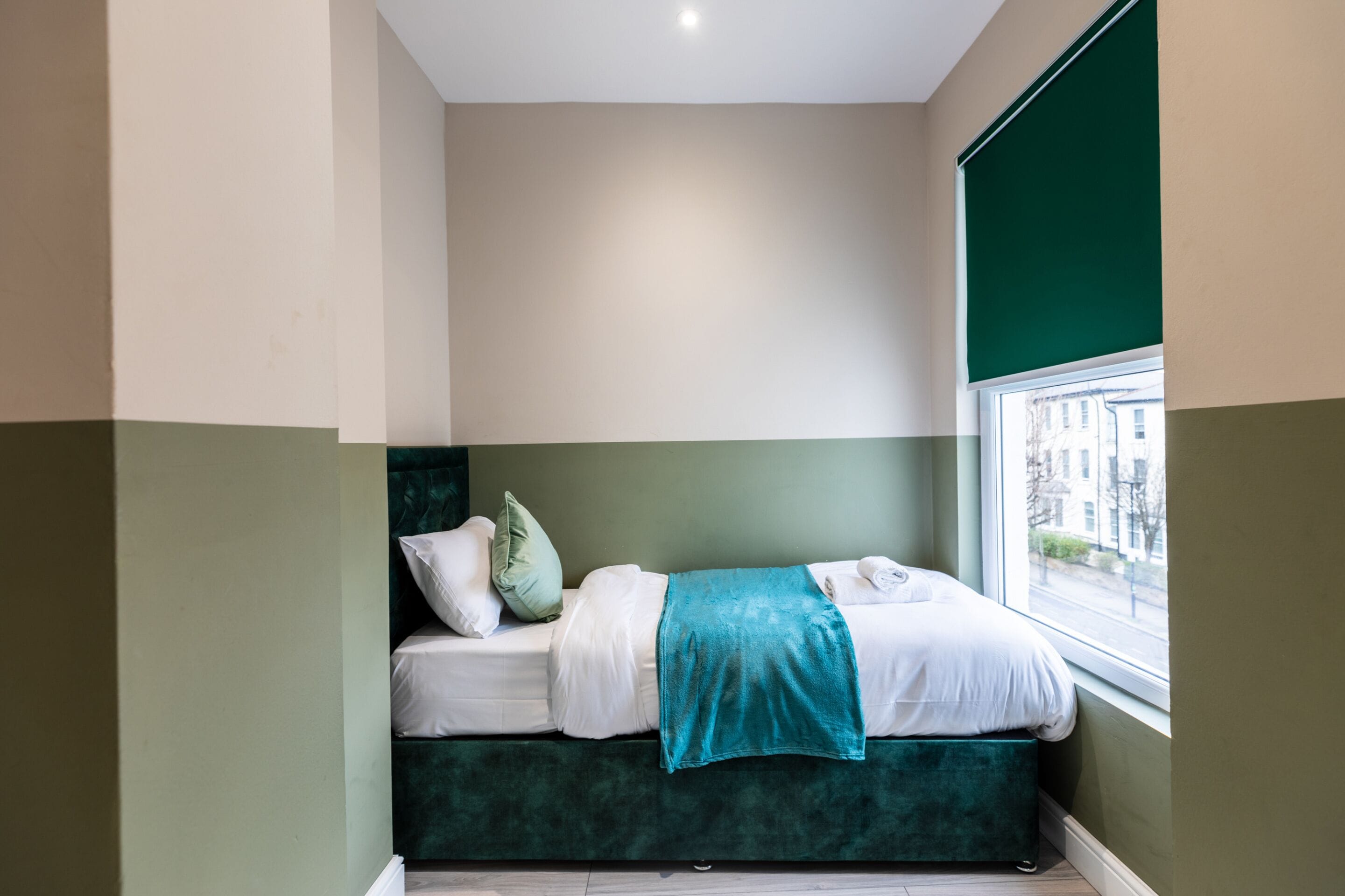 Un dormitorio pequeño y ordenado con una cama individual, una ventana con vistas a edificios y una pared de acento verde.