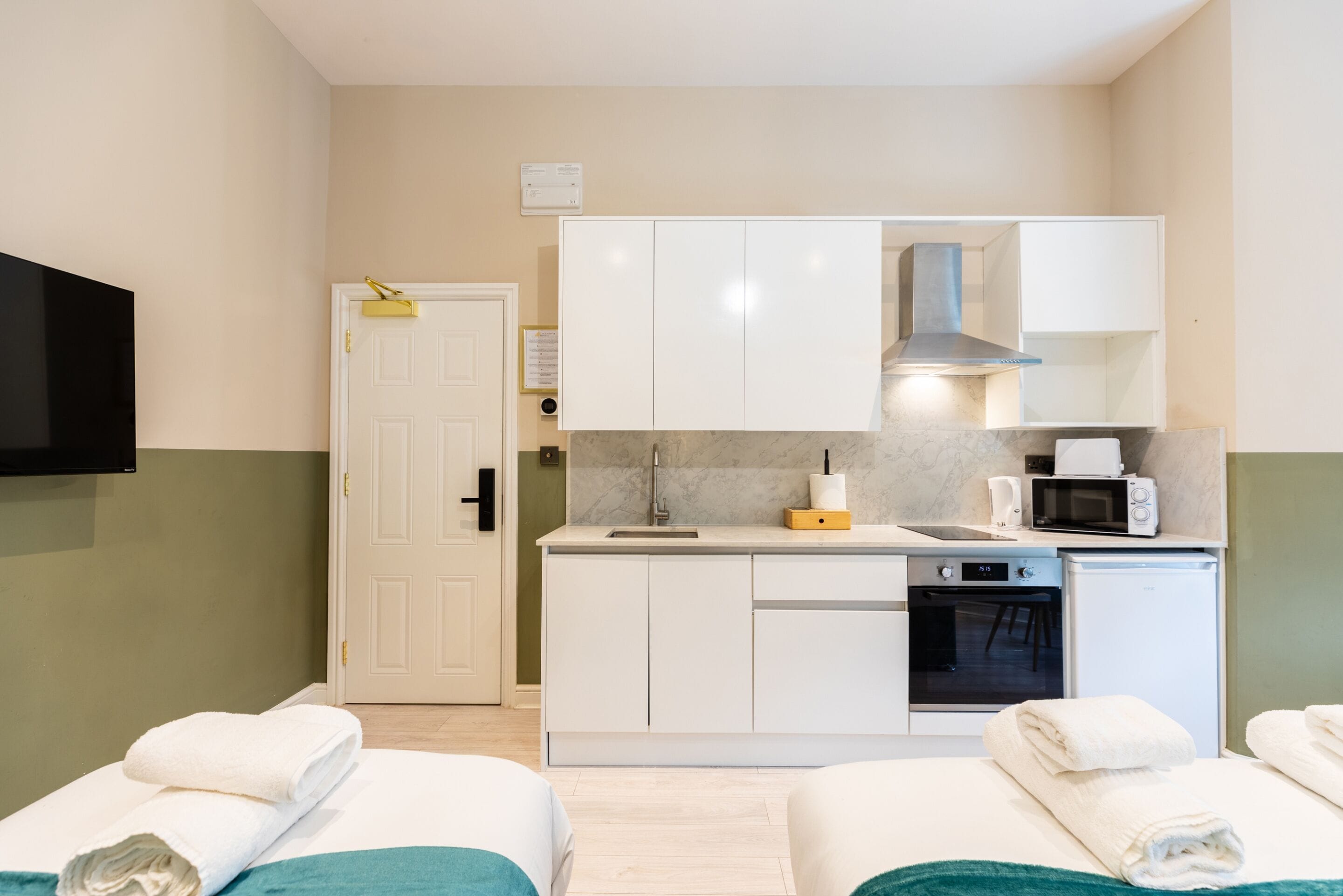 A compact hotel room with a kitchenette, a TV, and two beds with towels.