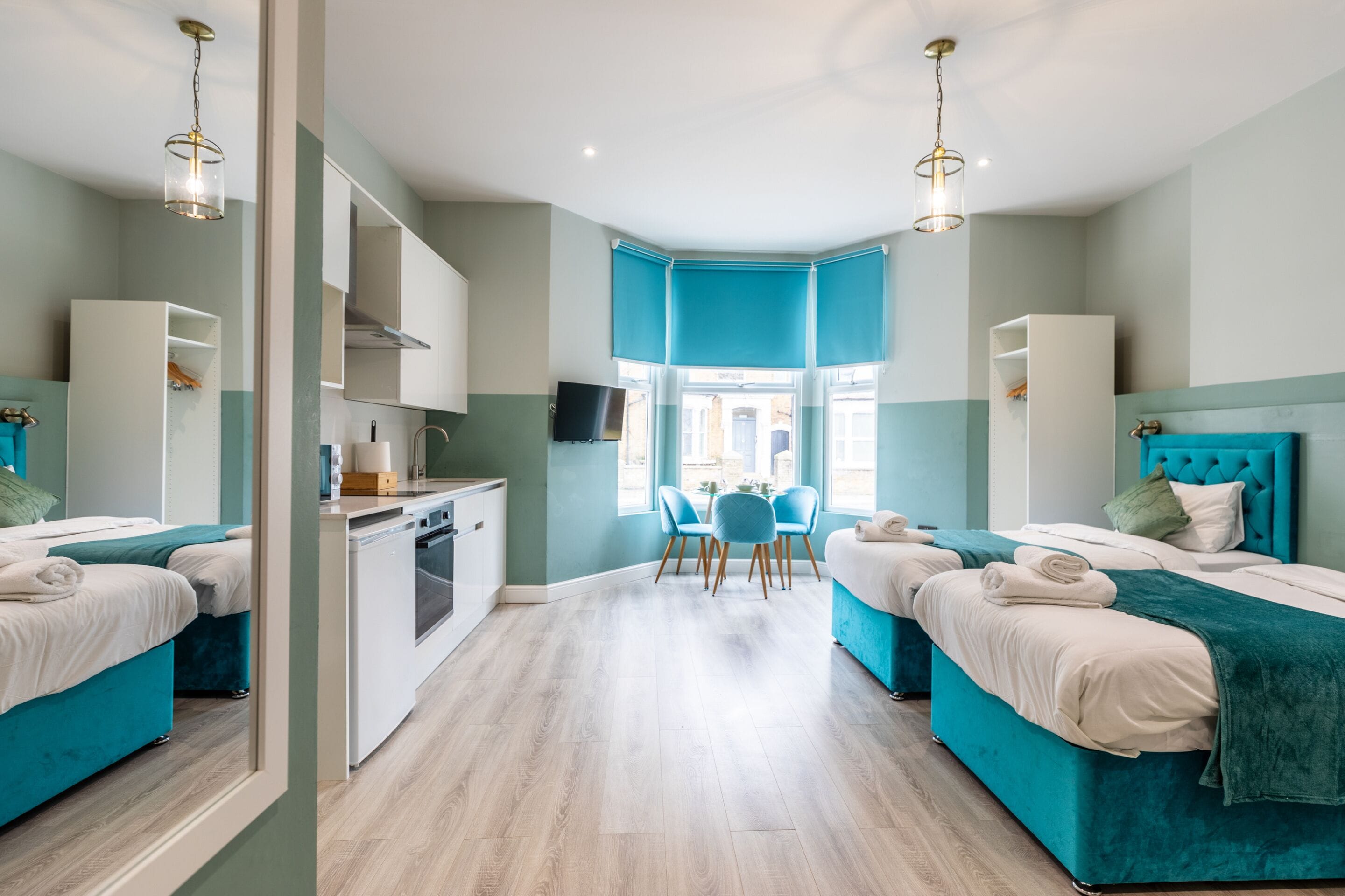 A modern hotel room with two beds, a dining area, and a kitchen.