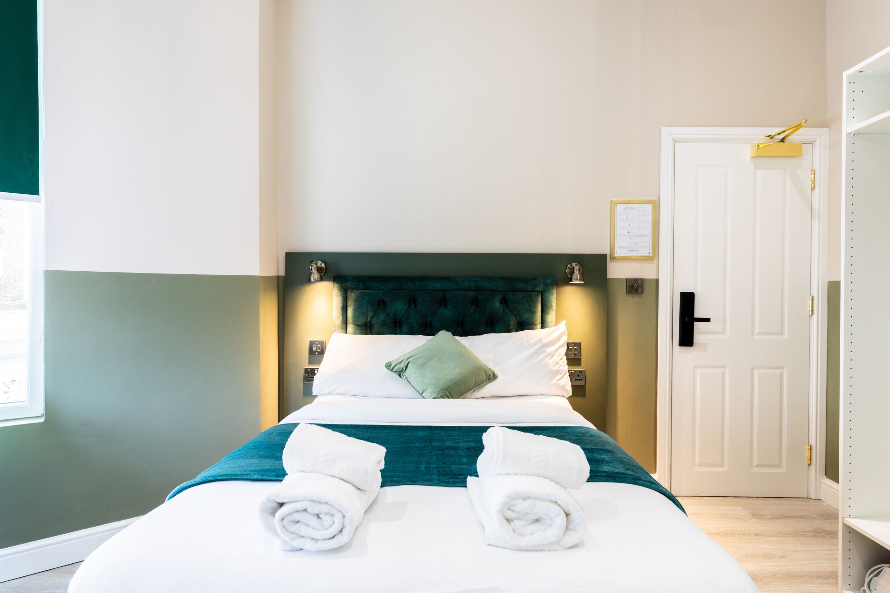 A neatly made bed with white linens and teal bedding, a green pillow, and two rolled towels on the bed.