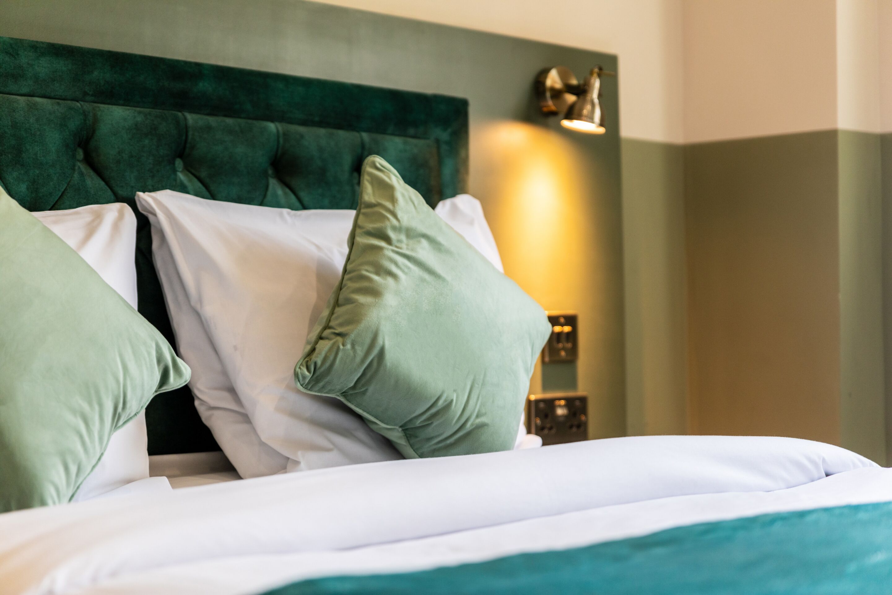 A neatly made bed with green and white pillows, a green upholstered headboard, and a wall-mounted light fixture.