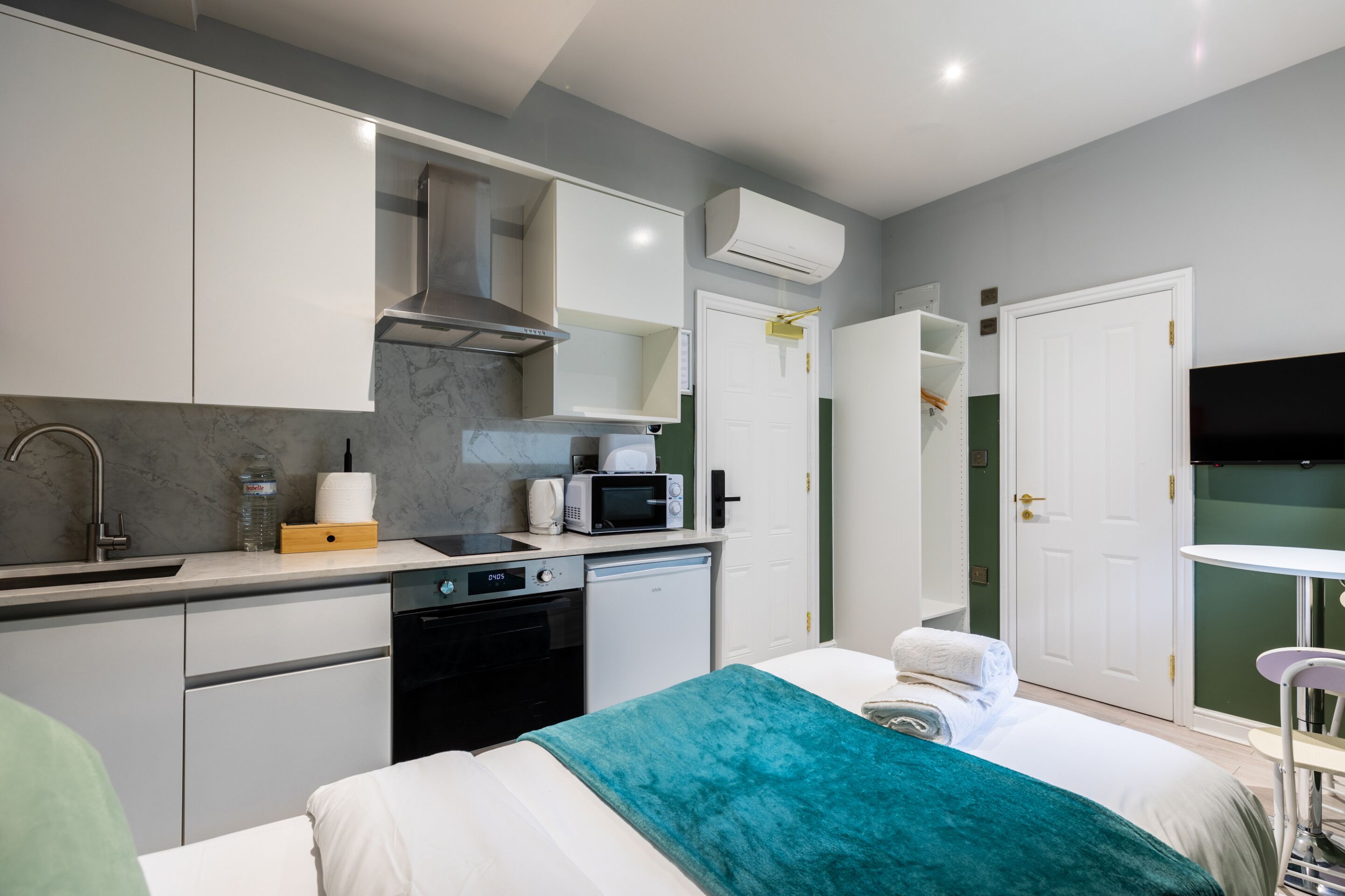A compact hotel room with a kitchenette, a bed with a teal blanket, a white door, and a small table with a chair.