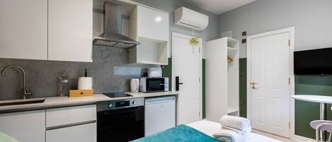 A compact hotel room with a kitchenette, a bed with a teal blanket, a white door, and a small table with a chair.