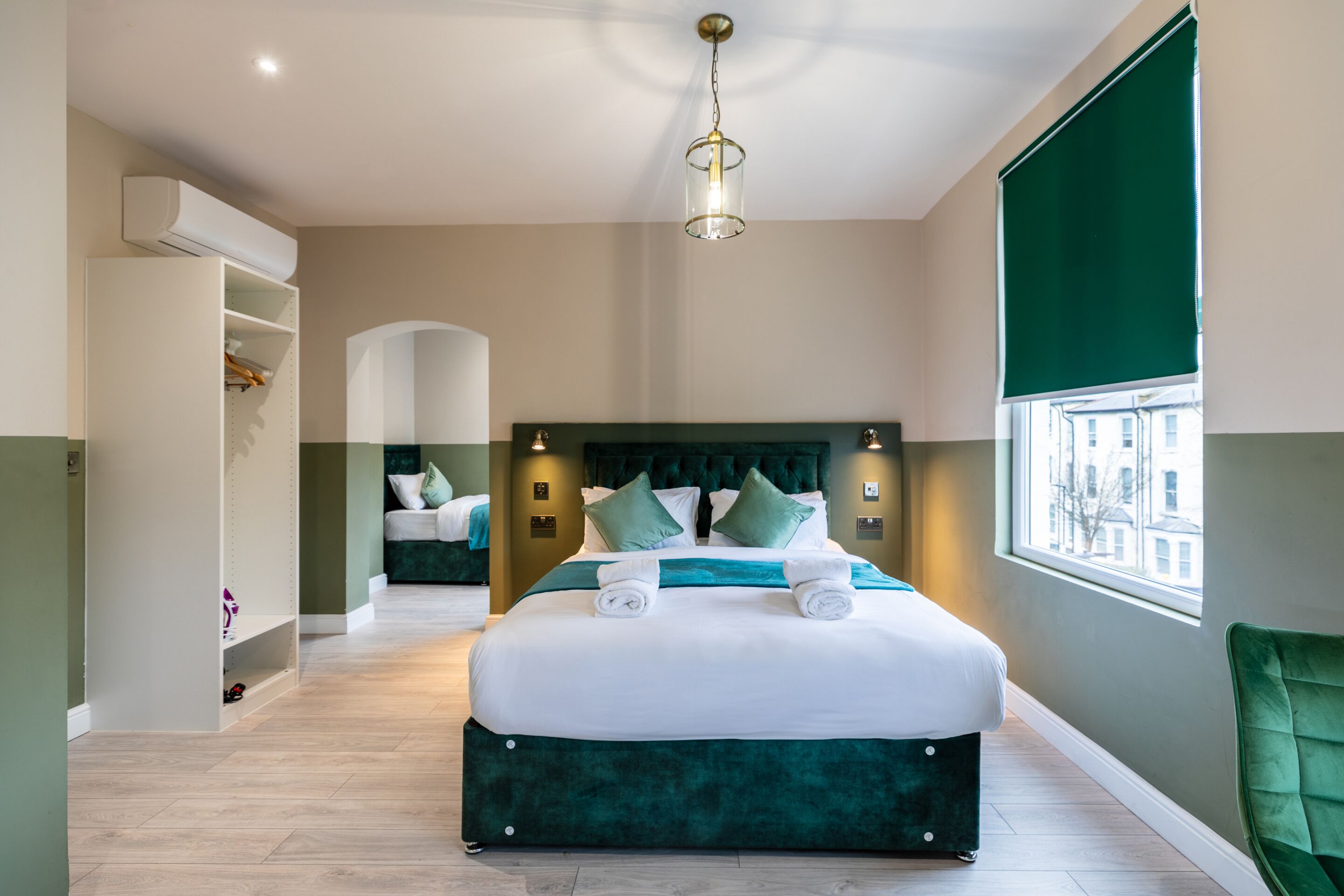 A modern hotel room with a large bed, a green upholstered headboard, a window with a view, and a built-in wardrobe.