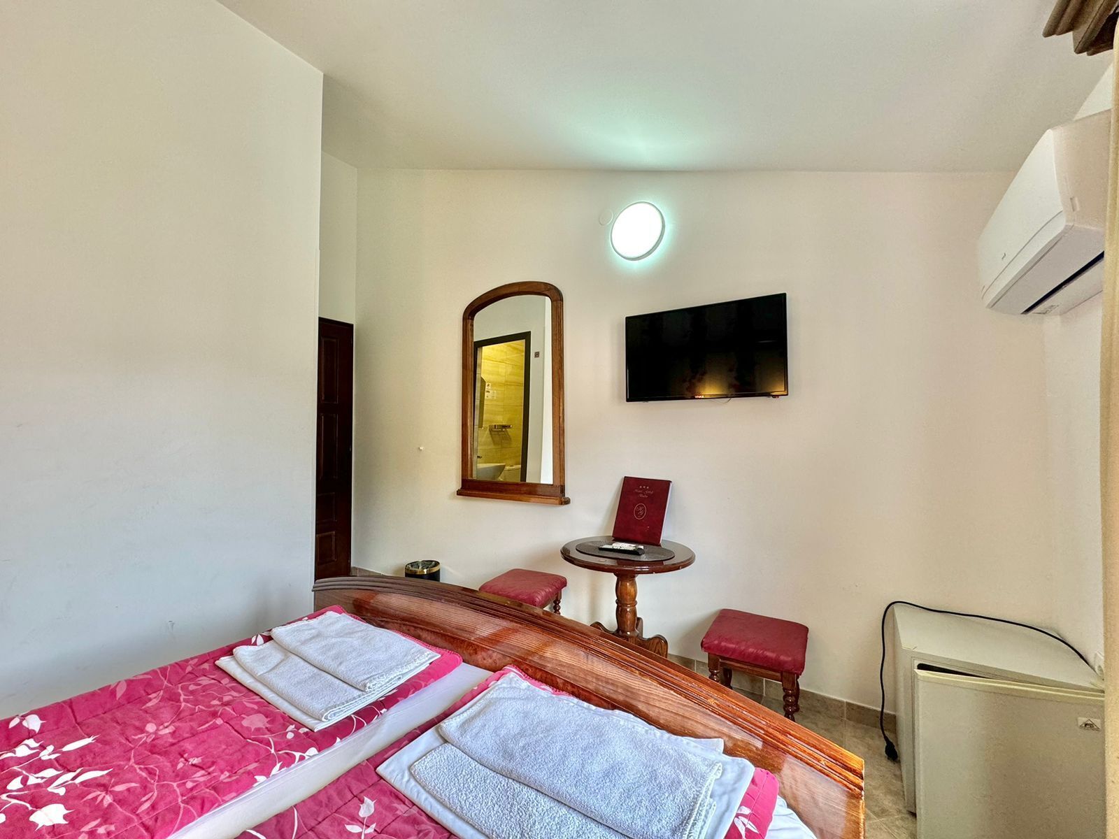 A hotel room with a bed, a TV, a mirror, a small table, and a refrigerator.