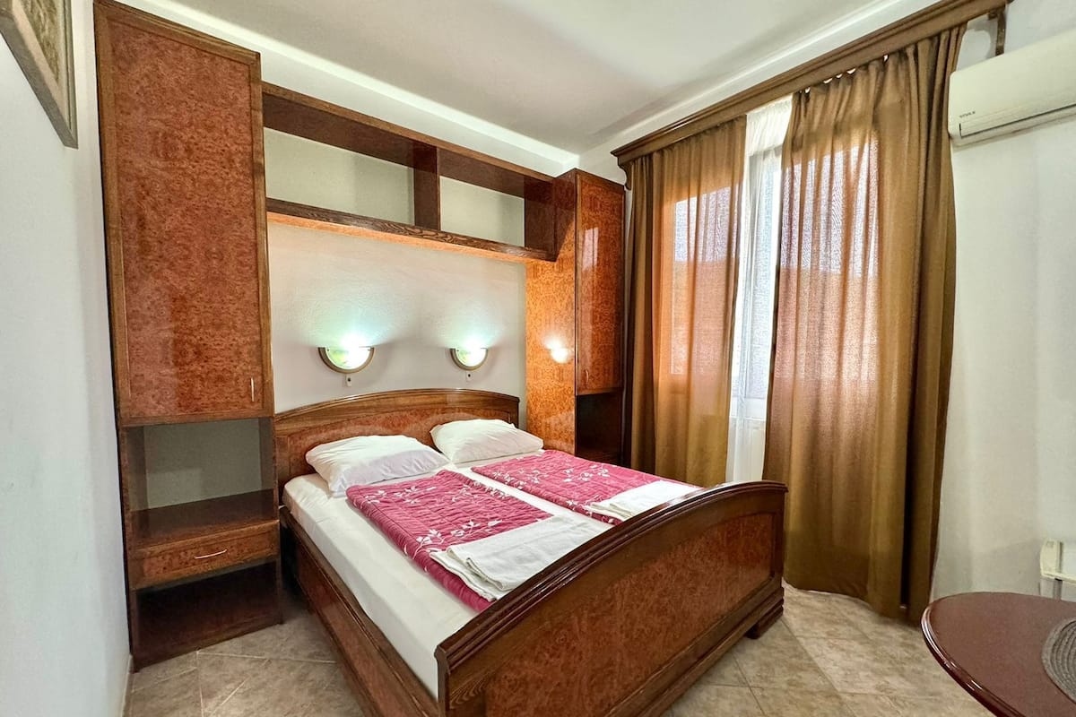 Une chambre d’hôtel avec un lit en bois, deux lampes de chevet, une étagère intégrée et une fenêtre avec des rideaux.