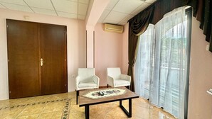 A room with a wooden door, two white armchairs, a small table with a placemat, a tiled floor, and a window with sheer curtains. - Hotel Grbalj (Budva)