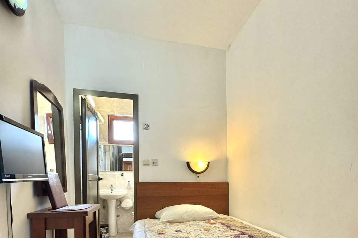 Une chambre d’hôtel comprenant un lit, un bureau avec un ordinateur, une télévision, un miroir, une lampe fixée au mur et une salle de bain visible à travers une porte ouverte.