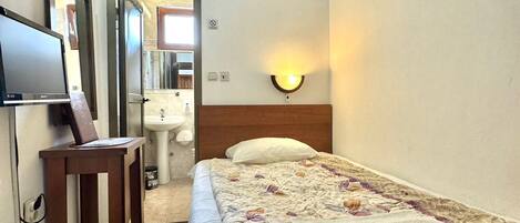Habitación de hotel con cama, escritorio con computadora, televisor, espejo, luz adherida a la pared y baño visible a través de una puerta abierta.
