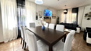 A modern living room with a dining area, featuring a dark wooden table, striped chairs, a blue vase with flowers, a flat-screen TV, and a black sofa. - STAY Villa Princess (Protaras)