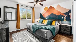 Une chambre à coucher comprenant un lit, un ventilateur de plafond, une table de chevet, une commode et une grande fenêtre avec des rideaux.