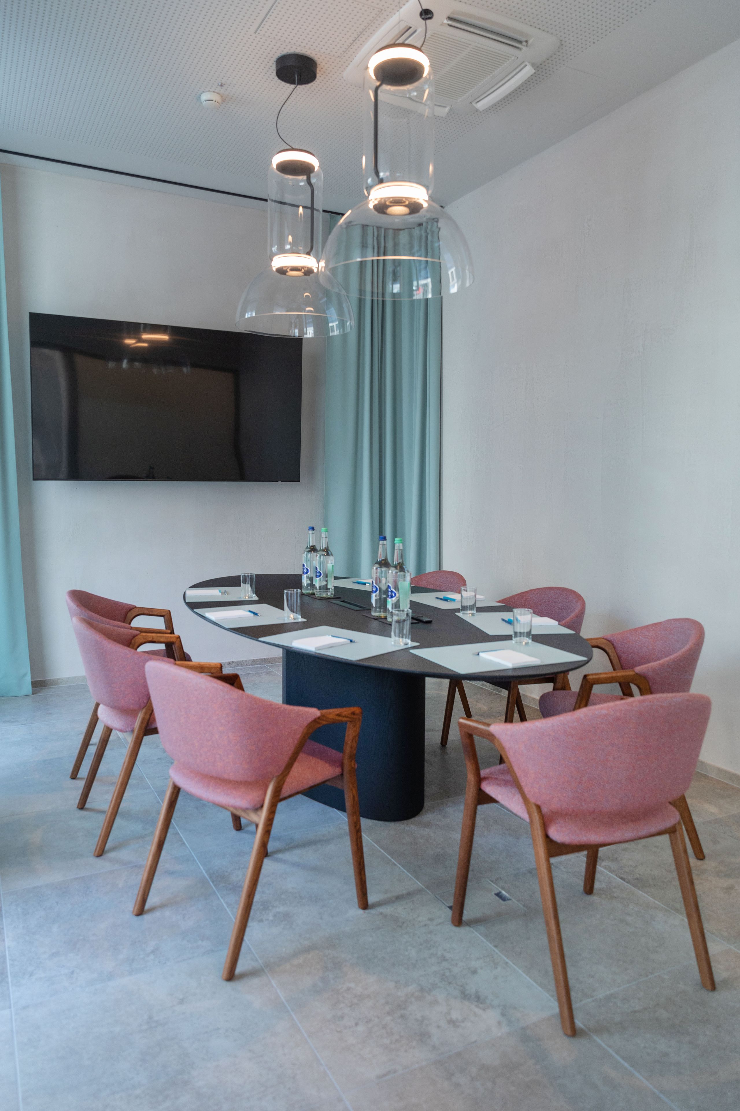 Une salle de conférence moderne, dotée d’une table ronde, de chaises rembourrées de couleur rose et d’un grand téléviseur à écran plat fixé au mur.
