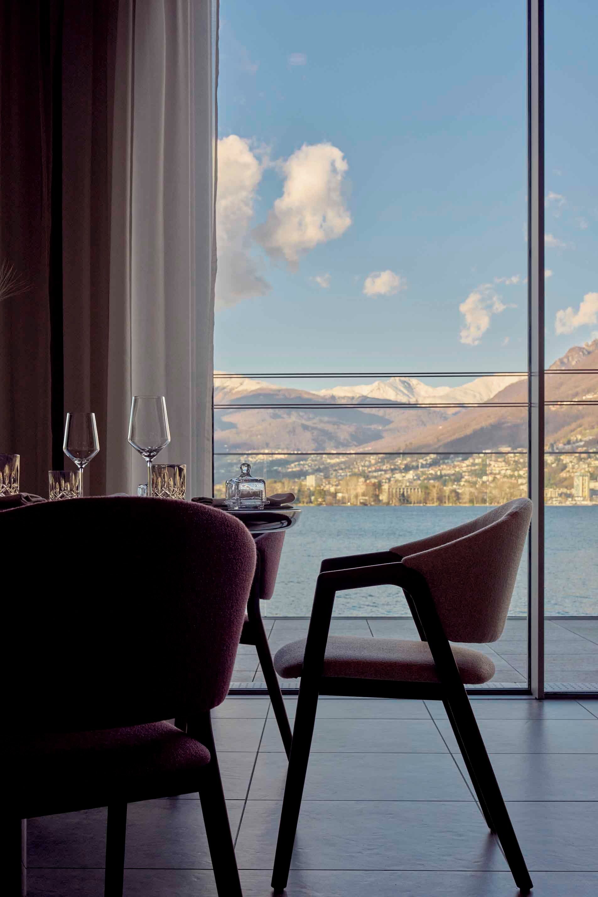 Un comedor con una mesa dispuesta para dos, una silla y vistas a una ciudad y montañas a través de grandes ventanales.