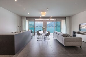A modern kitchen with a dining area, large windows with a view of the sea, and a living area with a sofa and TV. - Residenza Du Lac - Serviced Apartments (Paradiso)