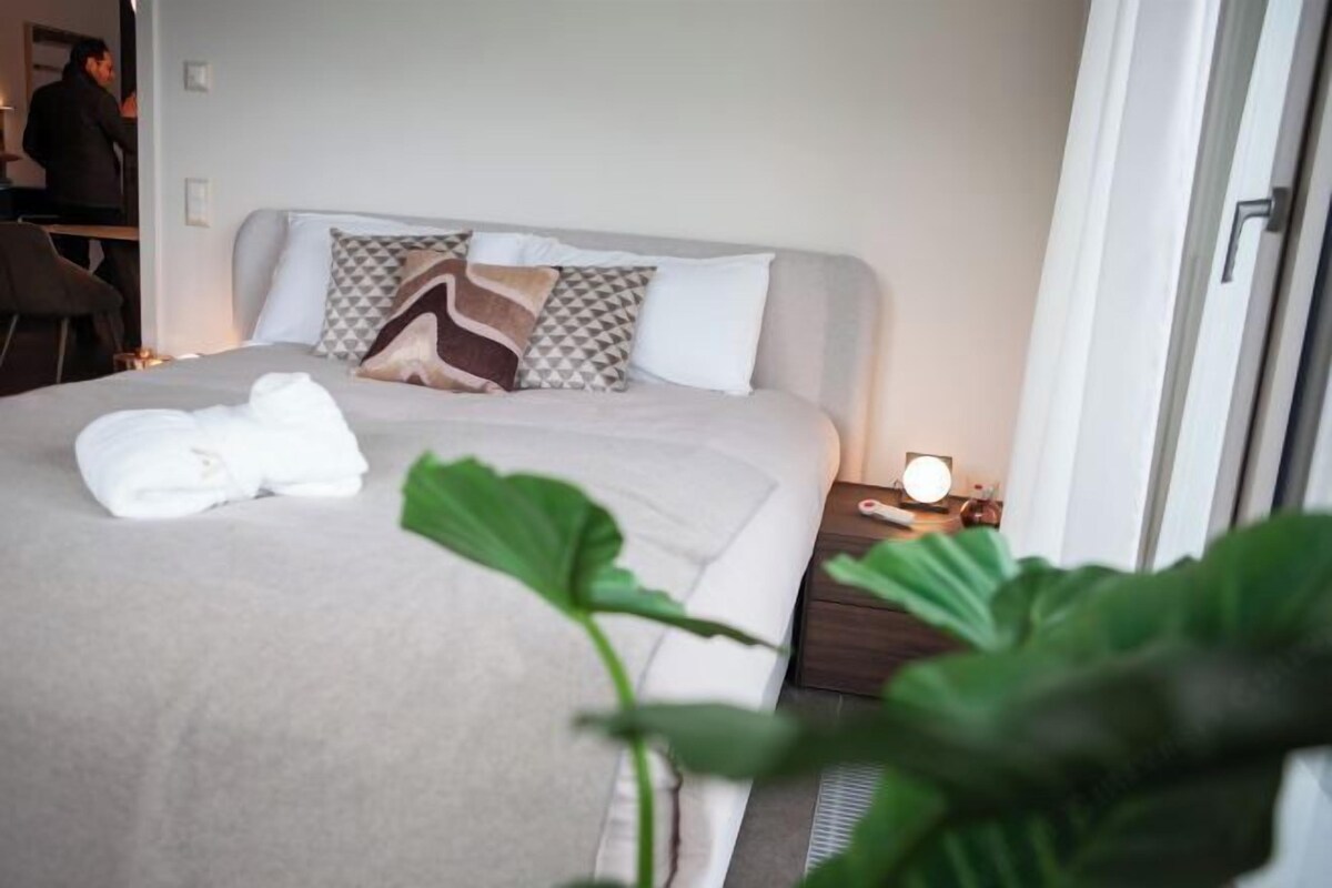 A neatly made bed with a white blanket and a towel, a bedside table with a lamp, and a large green plant in the foreground.