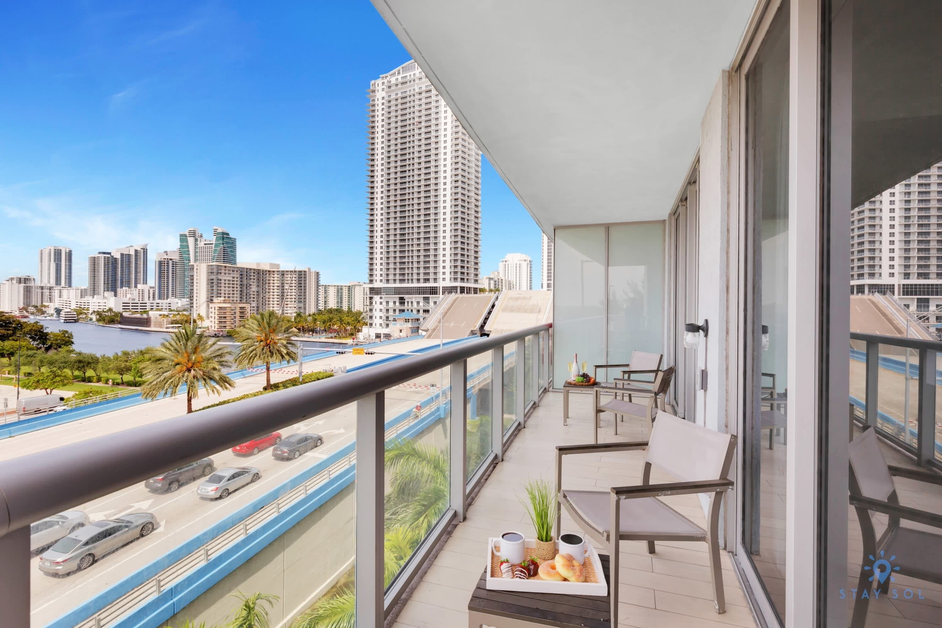 Un balcon avec vue sur la ville, un plateau avec des boissons et des en-cas, et un bâtiment moderne en arrière-plan.