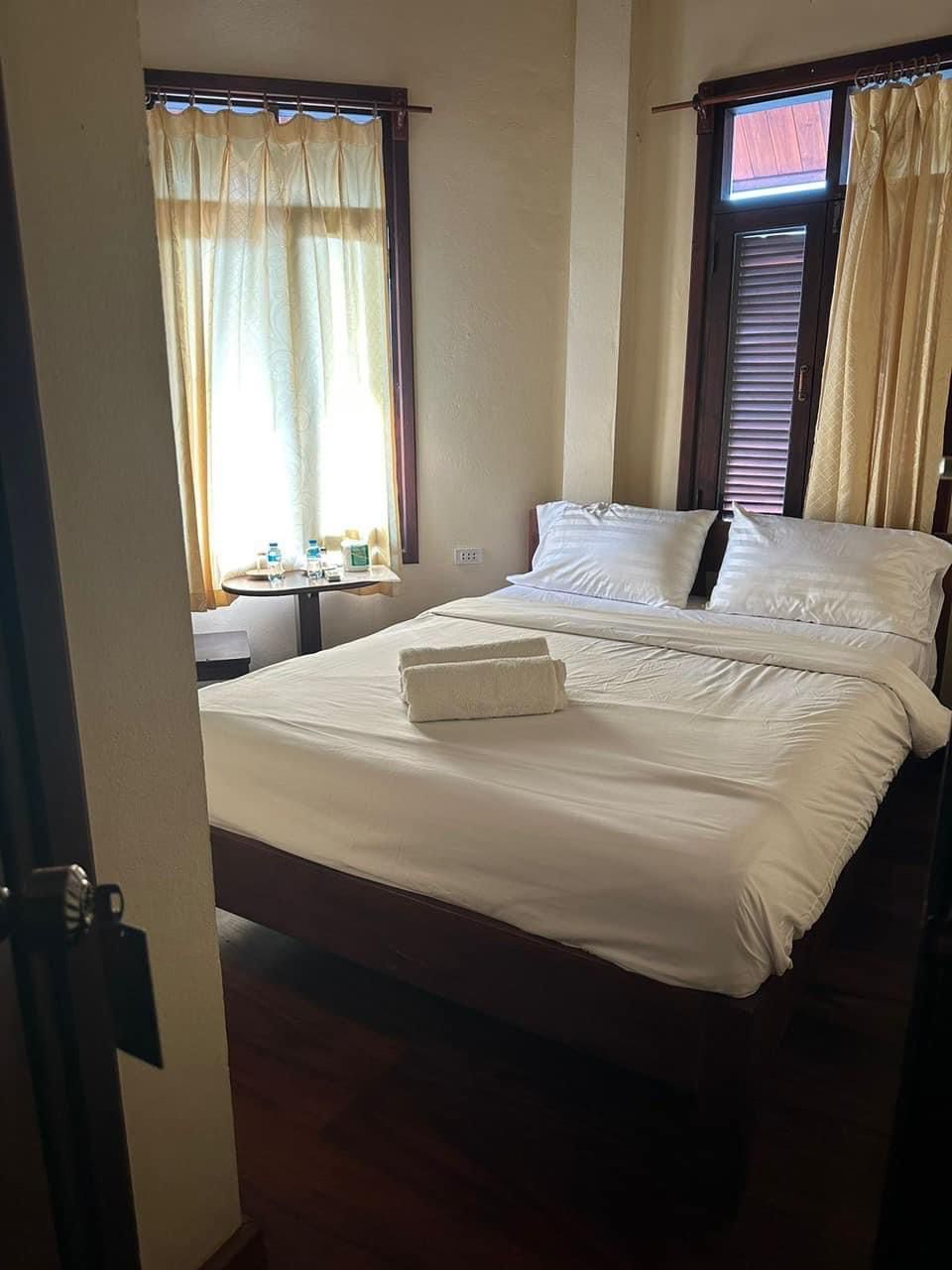 A hotel room with a bed, two windows with curtains, a bedside table with a bottle, and a towel on the bed.