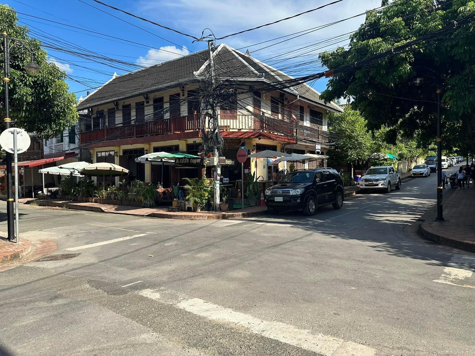 A street view with a two-story building featuring outdoor seating and umbrellas, a crosswalk, and parked cars.