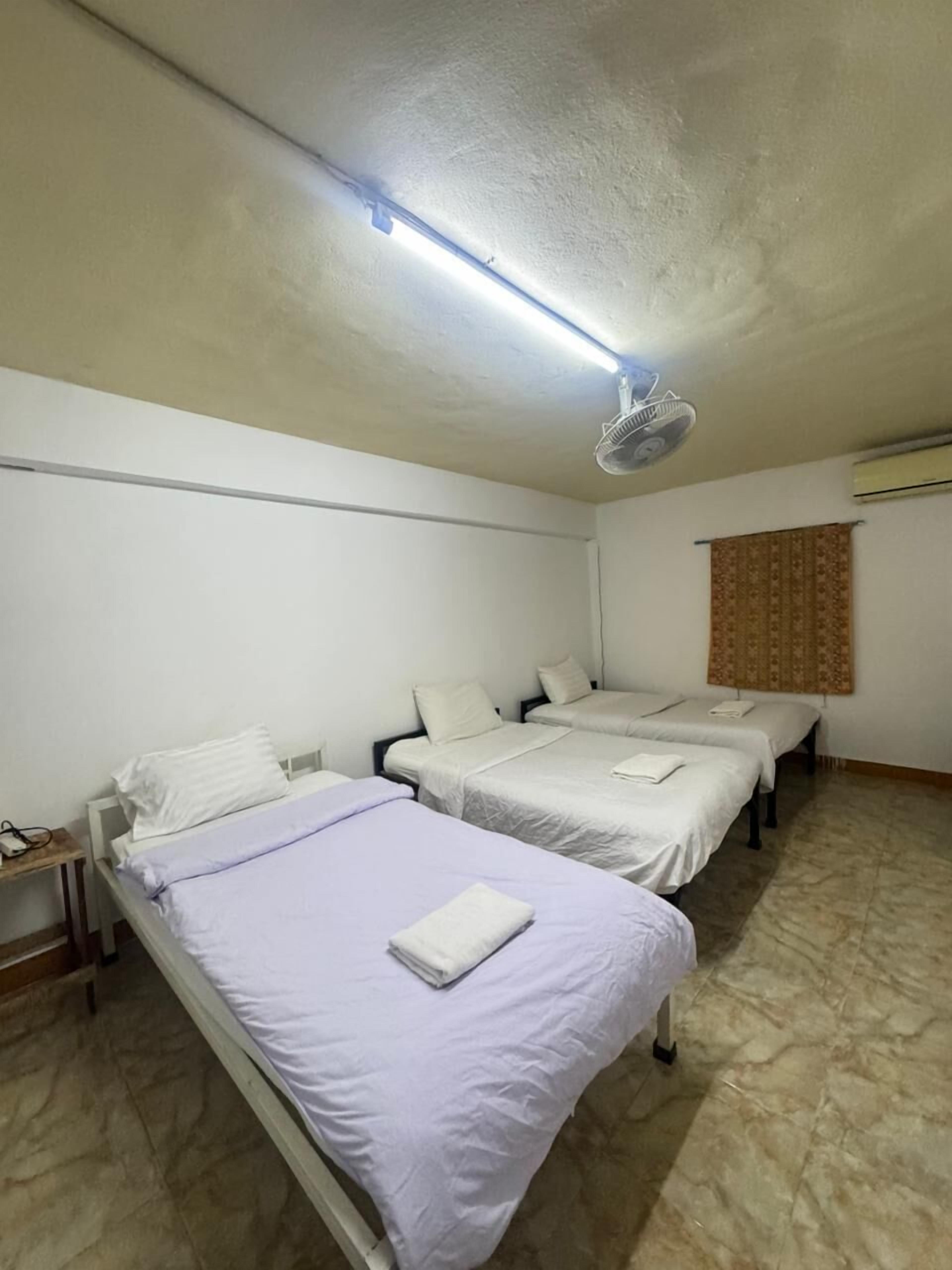 Ein Zimmer mit drei Betten, einem Deckenventilator, einer Lampe und einem Fenster mit Vorhängen.