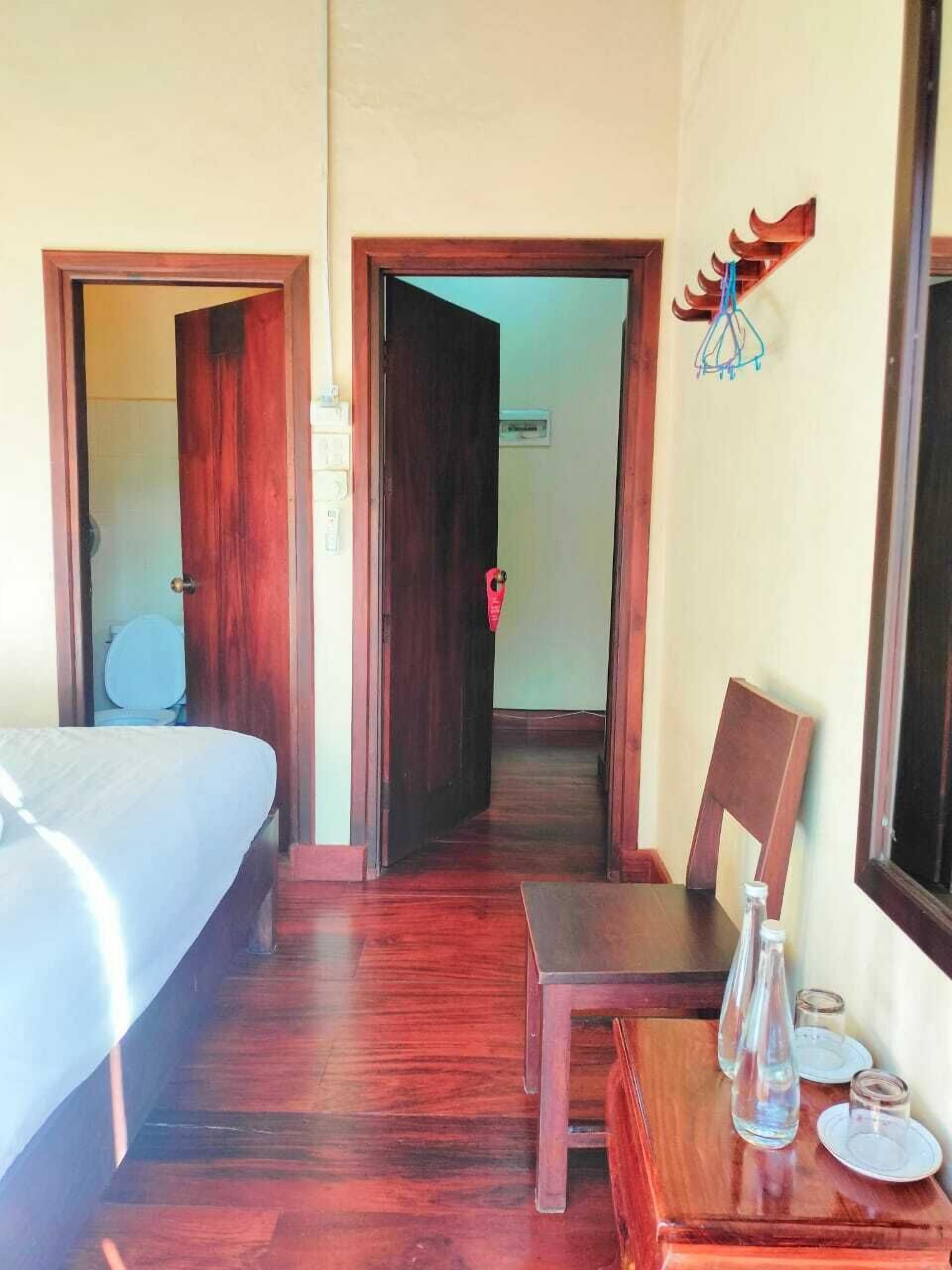 Habitación de hotel con una cama, una silla de madera, una mesita con vasos y una botella, y una puerta que da a otra habitación.