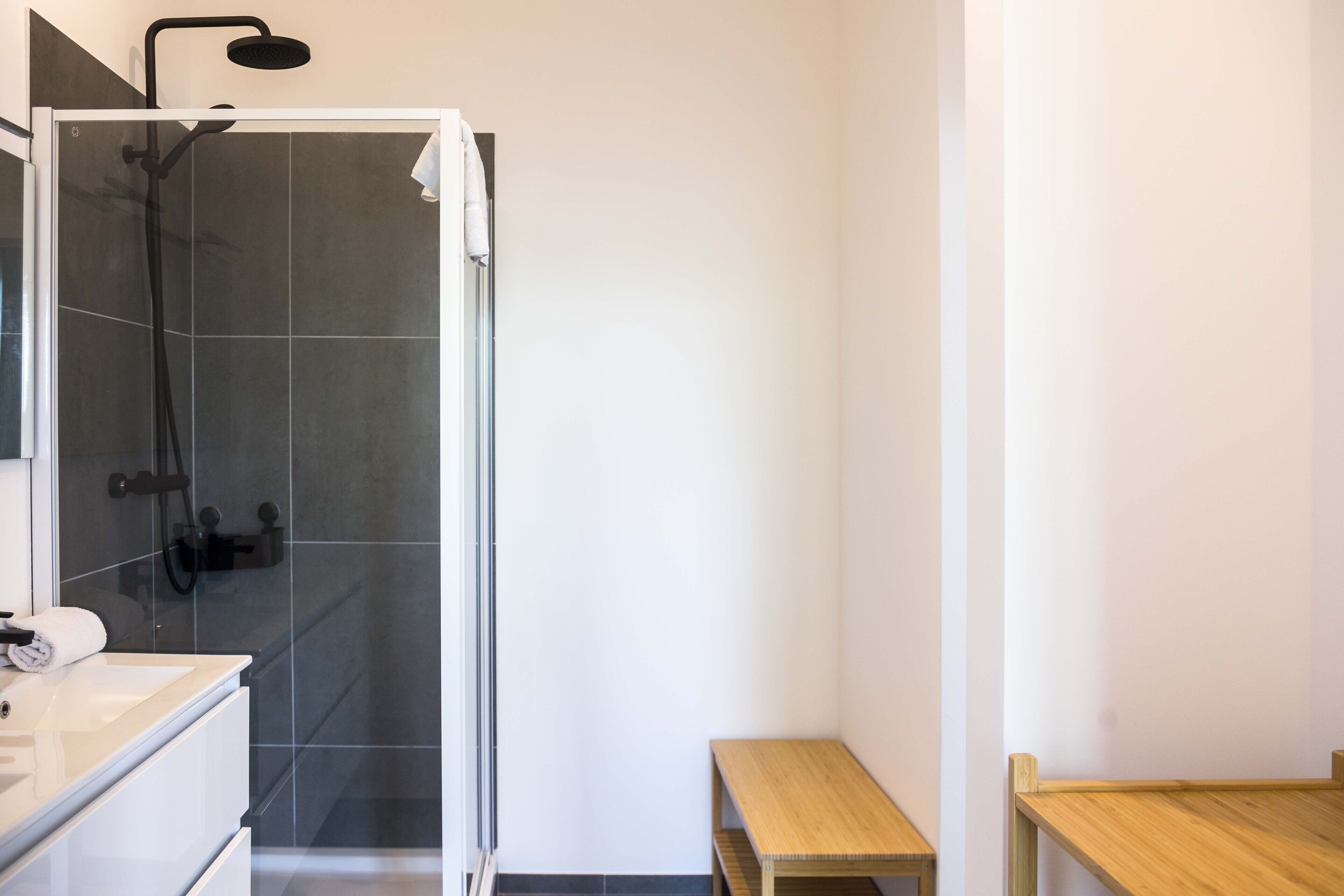 A modern bathroom with a glass-enclosed shower, a wooden bench, and a white vanity with a towel.