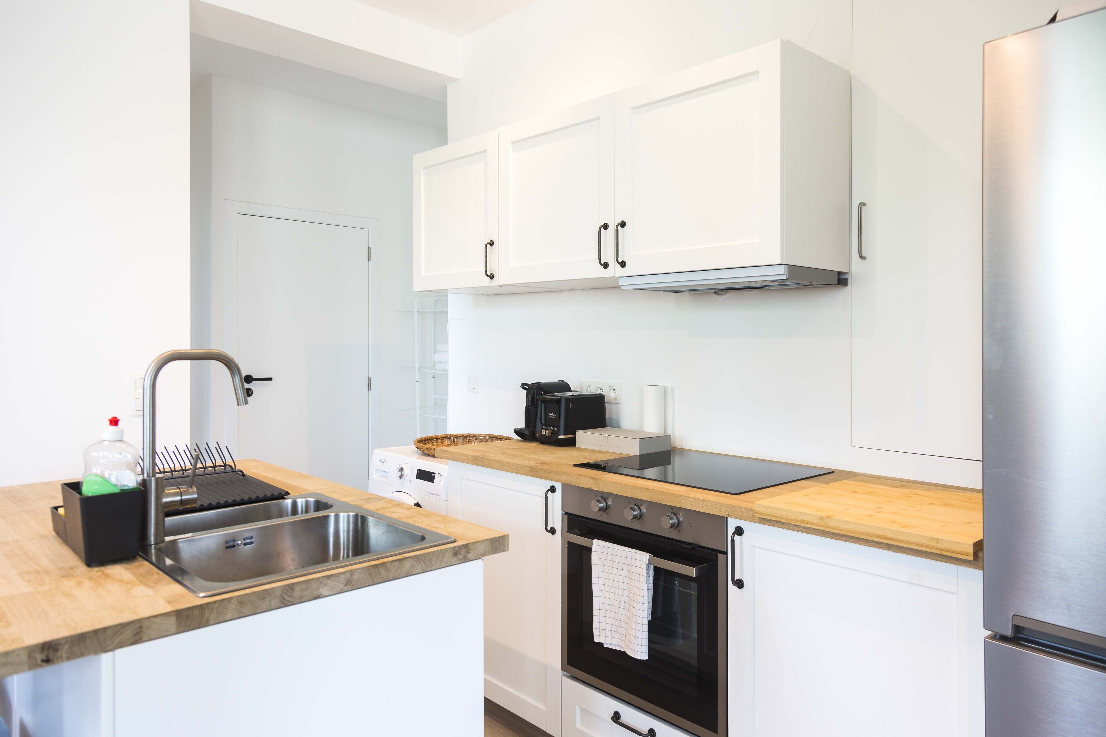 A modern kitchen with white cabinets, a stainless steel sink, a built-in oven, and a refrigerator.