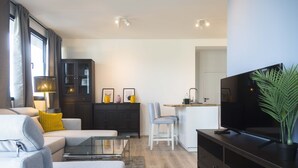Una sala de estar moderna con un sofá, una mesa de centro de vidrio, un televisor sobre un mueble bajo y una zona de cocina al fondo.