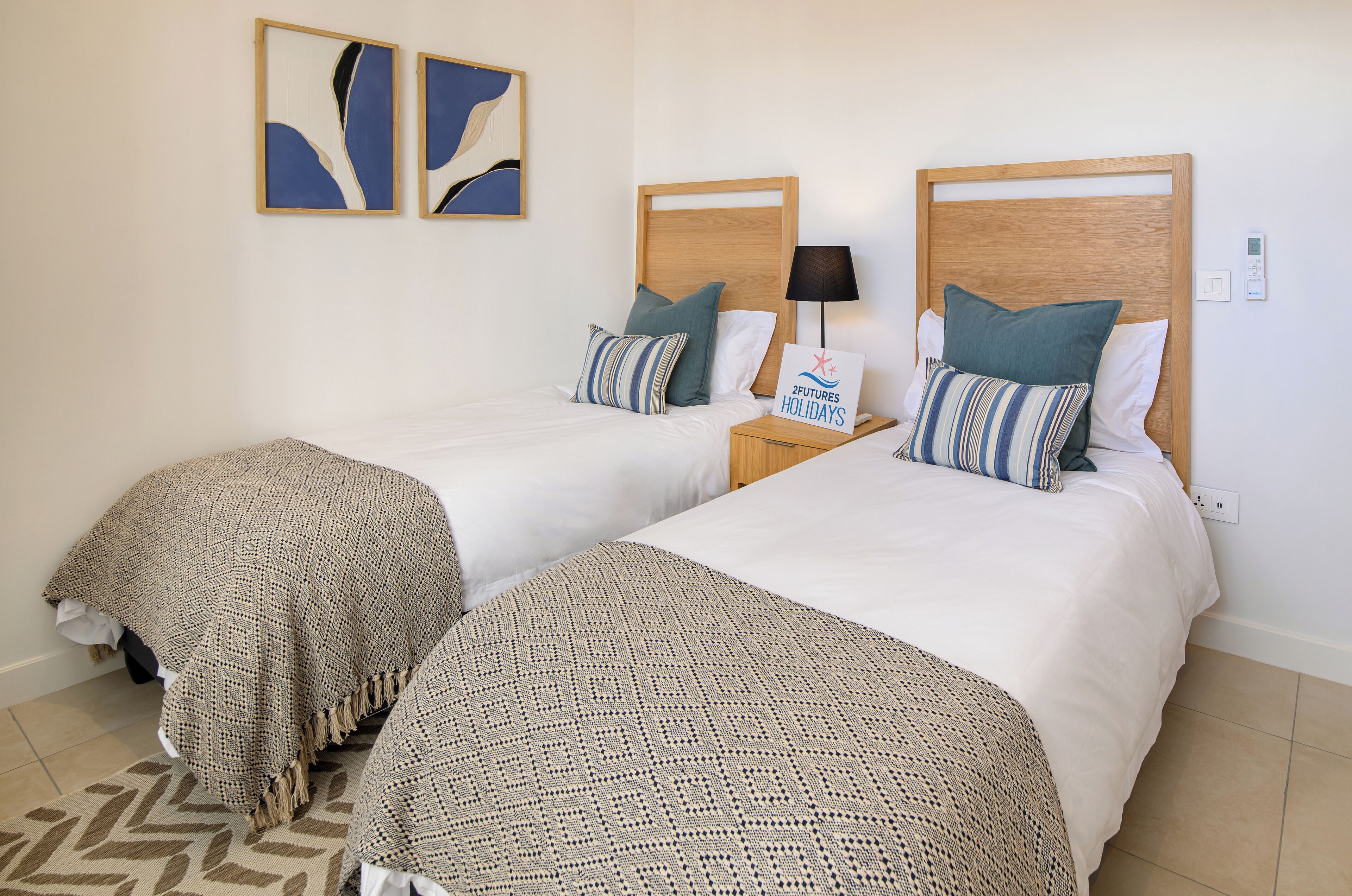 A hotel room with two beds, each with a wooden headboard, white bedding, and striped pillows. There is a nightstand with a lamp and a sign that reads "2Futures Holidays."