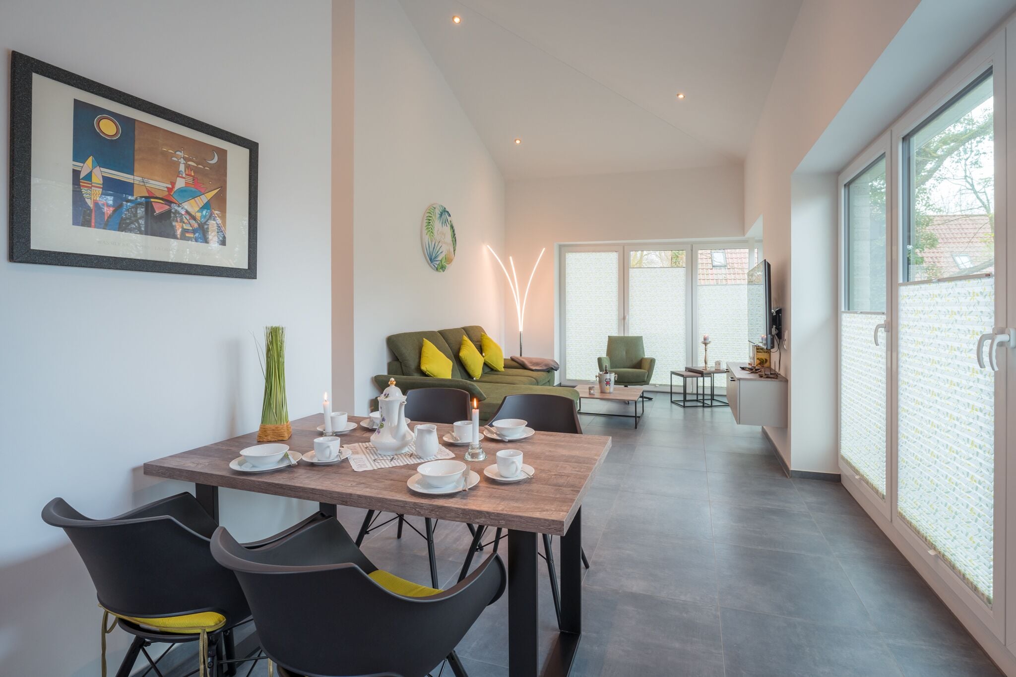 Un comedor moderno con una mesa de madera dispuesta para dos, un sofá verde con cojines amarillos, y un ventanal con vistas al exterior.