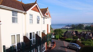A residential building with a balcony, a car parked in front, and a view of the sea and distant houses.