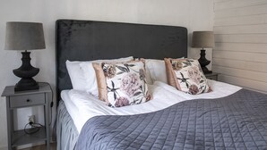 Een net opgemaakt bed met een grijze deken, twee bloemenkussens en een donker hoofdbord. Er staan twee nachtkastjes met lampen, en een daarvan heeft een stopcontact.