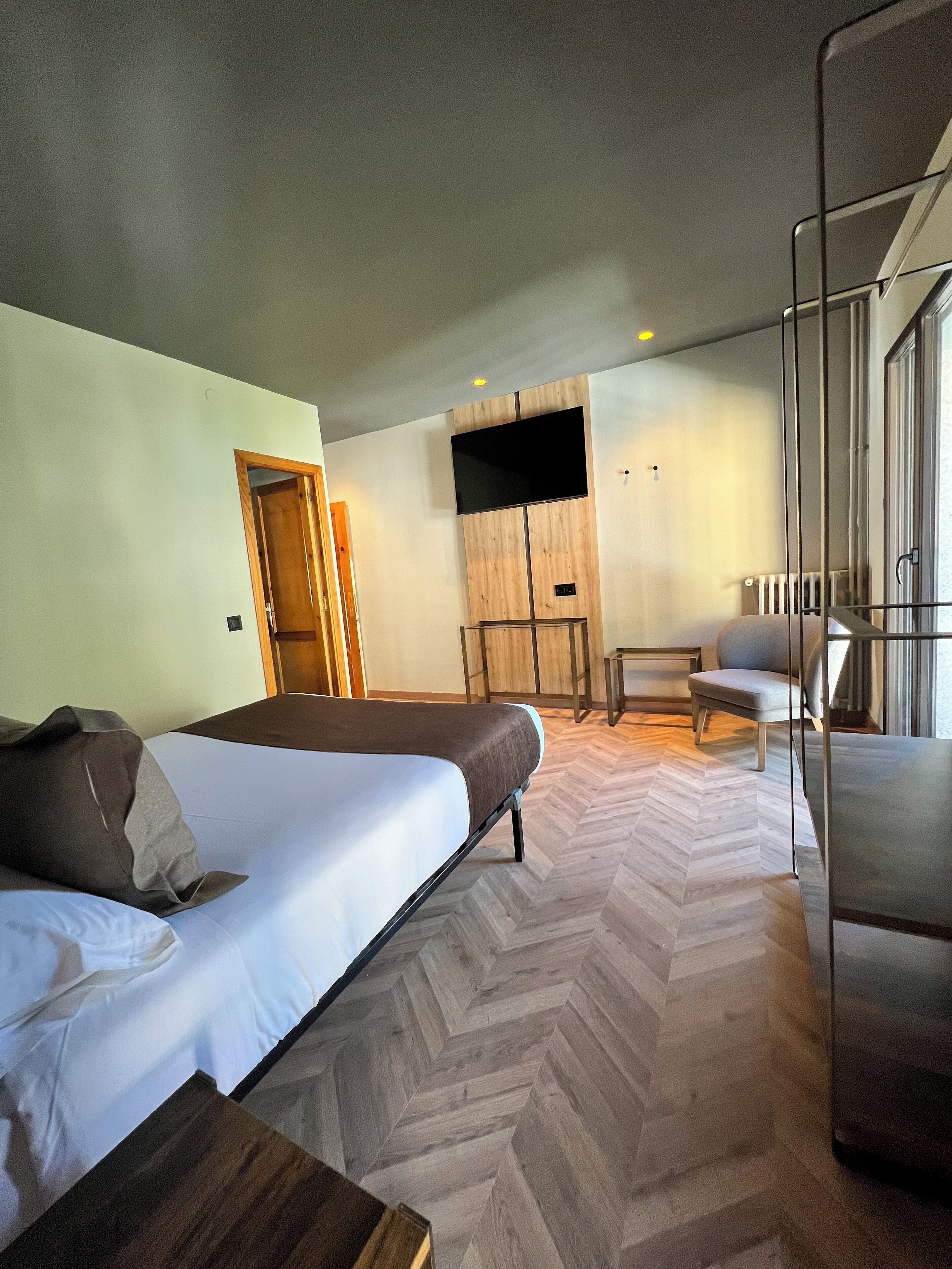 A modern hotel room with a large bed, a seating area with a chair, a TV mounted on a wooden panel, and a herringbone-patterned floor.