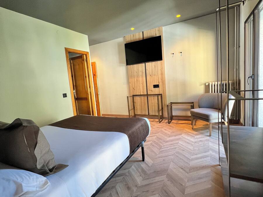 Una habitación de hotel moderna con una cama grande, un área de descanso con una silla, un televisor montado en un panel de madera y un suelo con estampado de espiga.