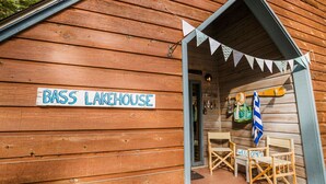 Eine Holzhütte mit dem Schild "Bass Lakehouse", inklusive einer kleinen Veranda mit Tisch und Stühlen sowie einem Schild mit der Aufschrift "Willkommen".