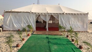 A large white tent with a red carpet entrance, surrounded by small palm trees and pots on a sandy beach. - Kumbh Village - Dining By Taj Ganges (Karchana)