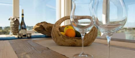 Two wine glasses on a wooden table with a basket of fruit and a bottle in the background.