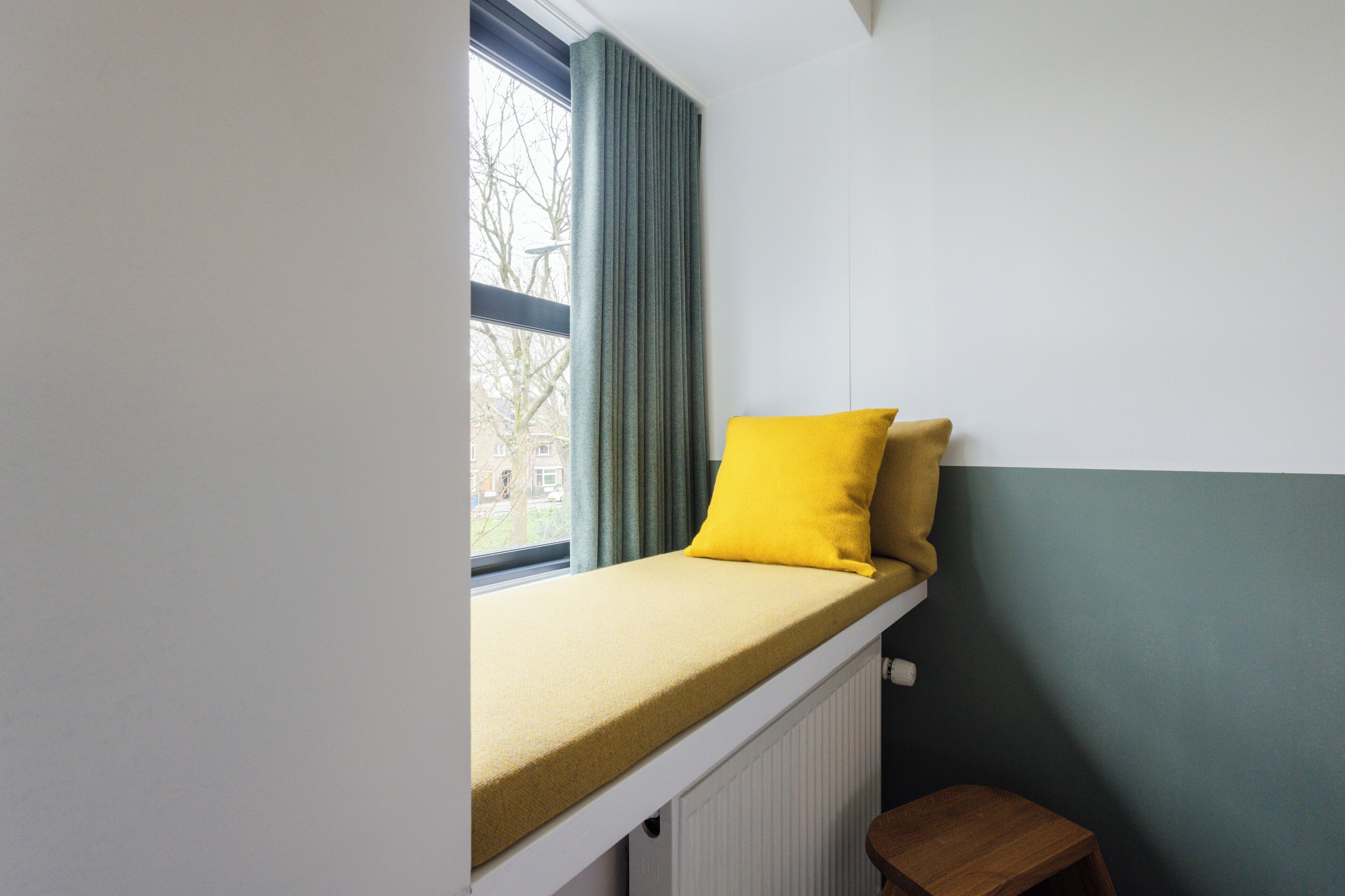 Een kamer met een raam, een radiator en een klein houten krukje.