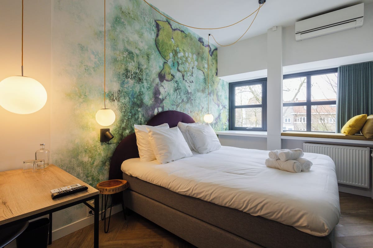 A hotel room with a bed, bedside table, lamp, and a view of the outside through a window.