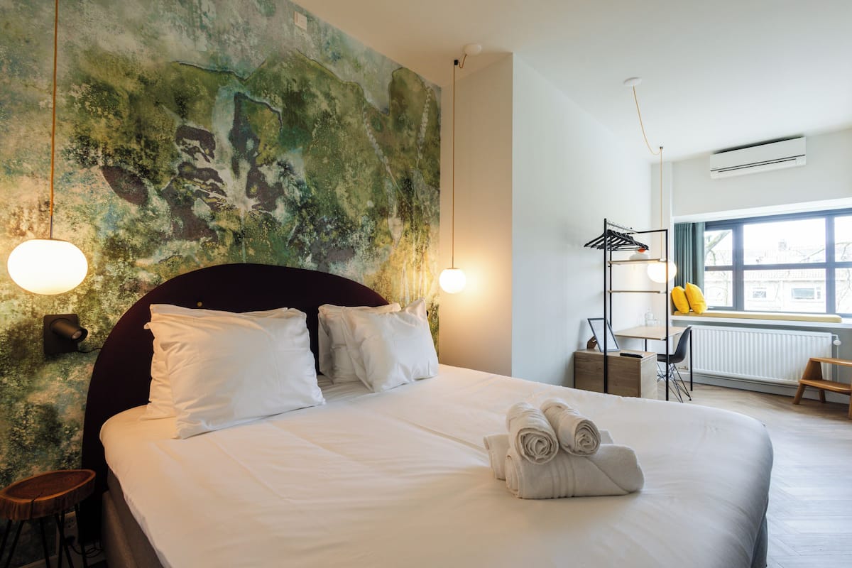 A hotel room with a large bed, a headboard, a wall-mounted shelf with towels, a small table, and a window with a view of the city.
