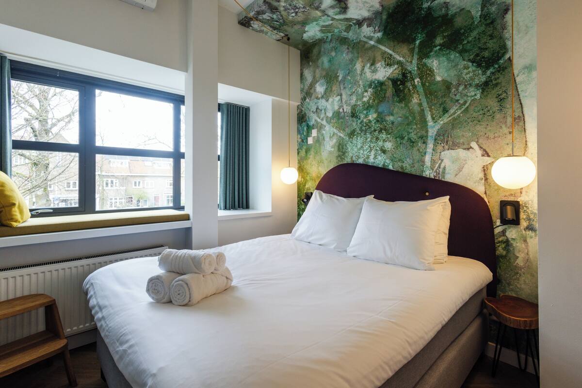 A neatly made bed with white linens and rolled towels, a window with a view of buildings, and a wall with a textured mural.